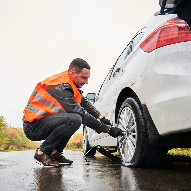Male auto mechanic in work vest unscrewing lug nuts on car wheel in process of new tire replacement. Young man using wrench while changing flat tire on the road. Concept of emergency road service.